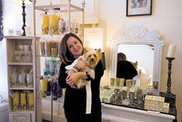 Shop owner is holding her dog. There are candles and cups in the displays behind her.